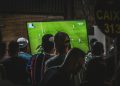 Grupo de personas de espaldas viendo la retransmisión de un partido de fútbol en una televisión de gran tamaño.