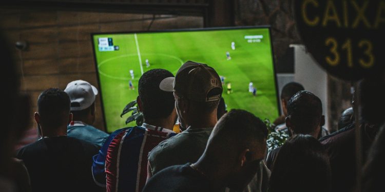 Grupo de personas de espaldas viendo la retransmisión de un partido de fútbol en una televisión de gran tamaño.