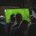 Grupo de personas de espaldas viendo la retransmisión de un partido de fútbol en una televisión de gran tamaño.